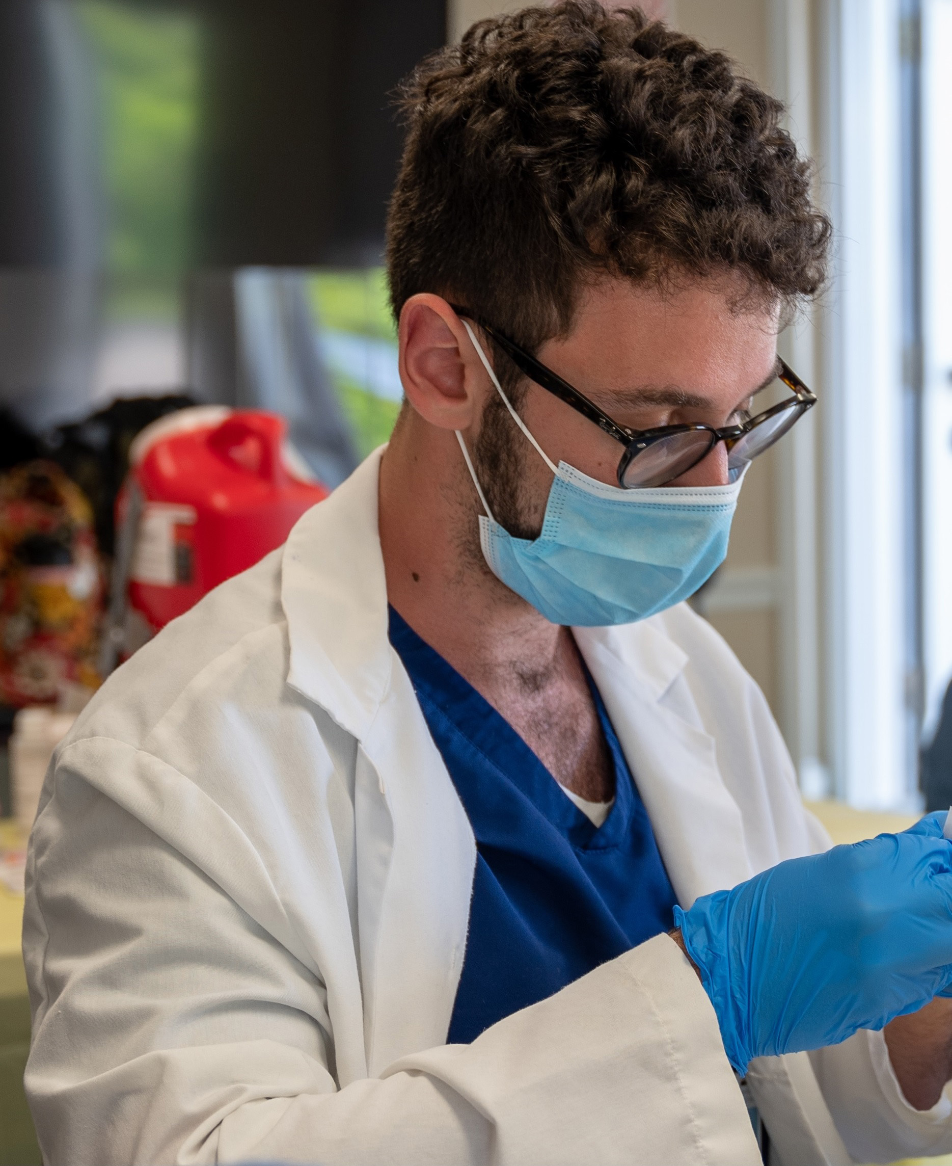 A male student wearing a surgical mask and latex gloves, blue scrubs, and a white lab coat looks down at an item he is holding